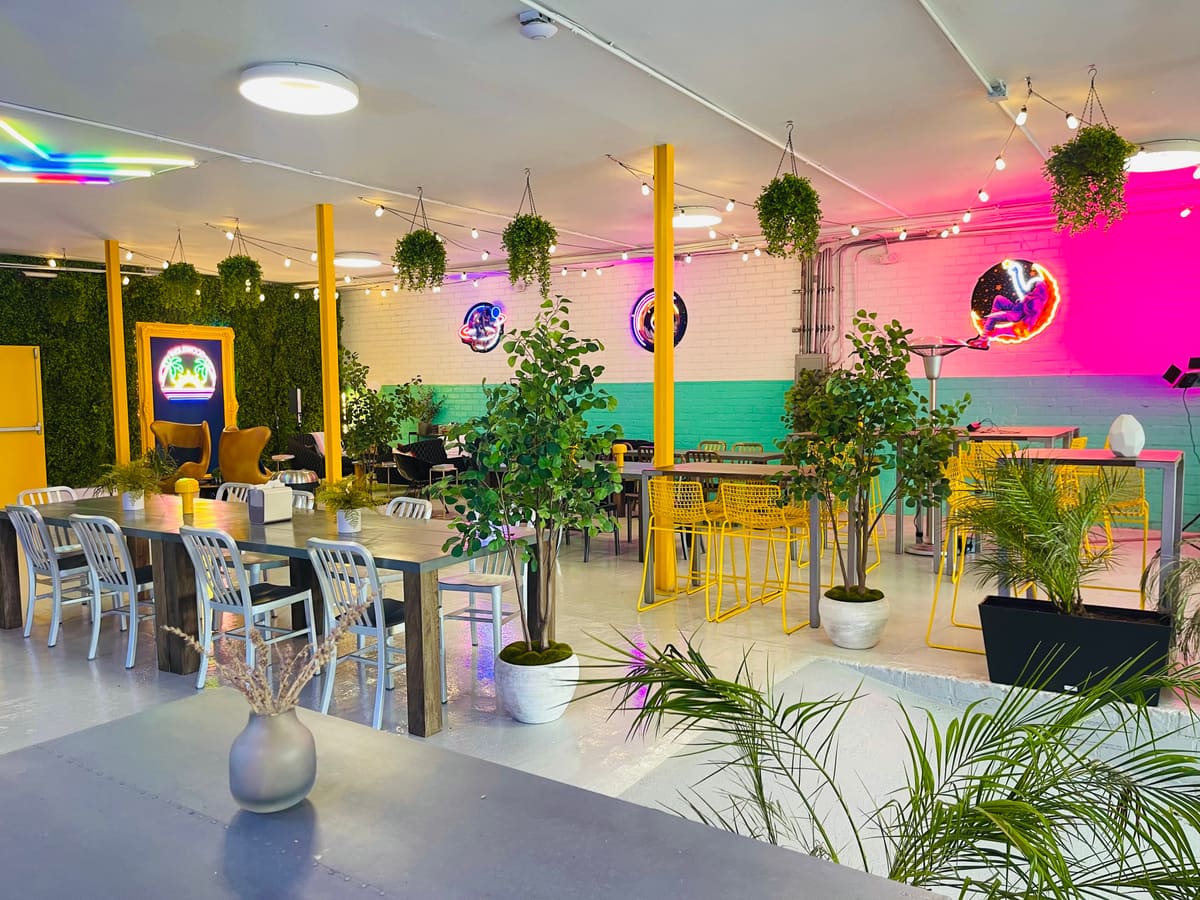 Café seating area with metal chairs, high-top tables, potted palms, string lights and colorful neon accent walls