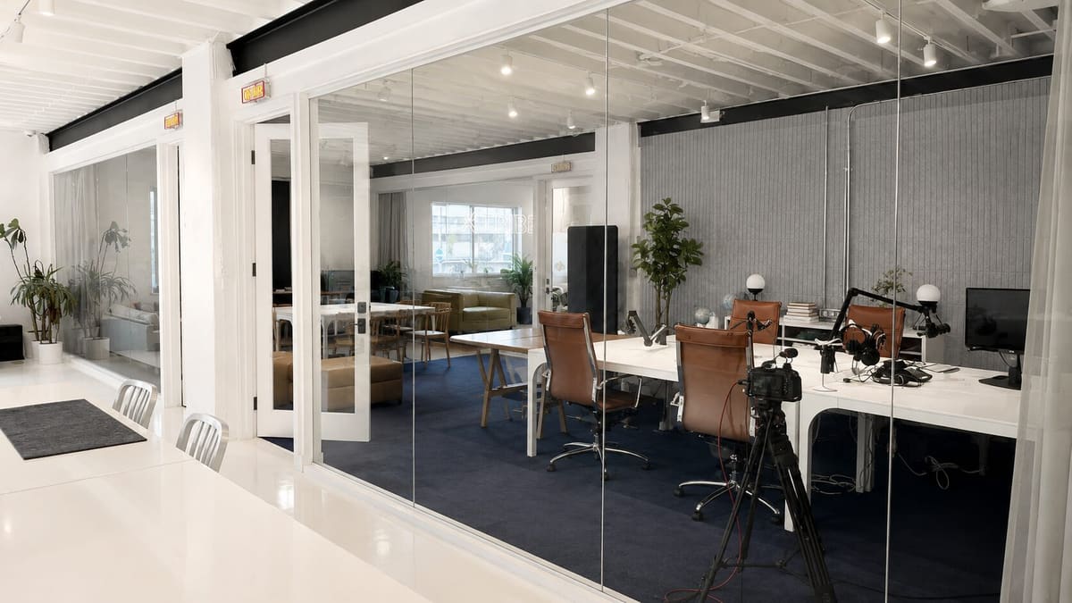 Glass-walled room with a white table, leather chairs and podcast microphones, seen from a bright hallway
