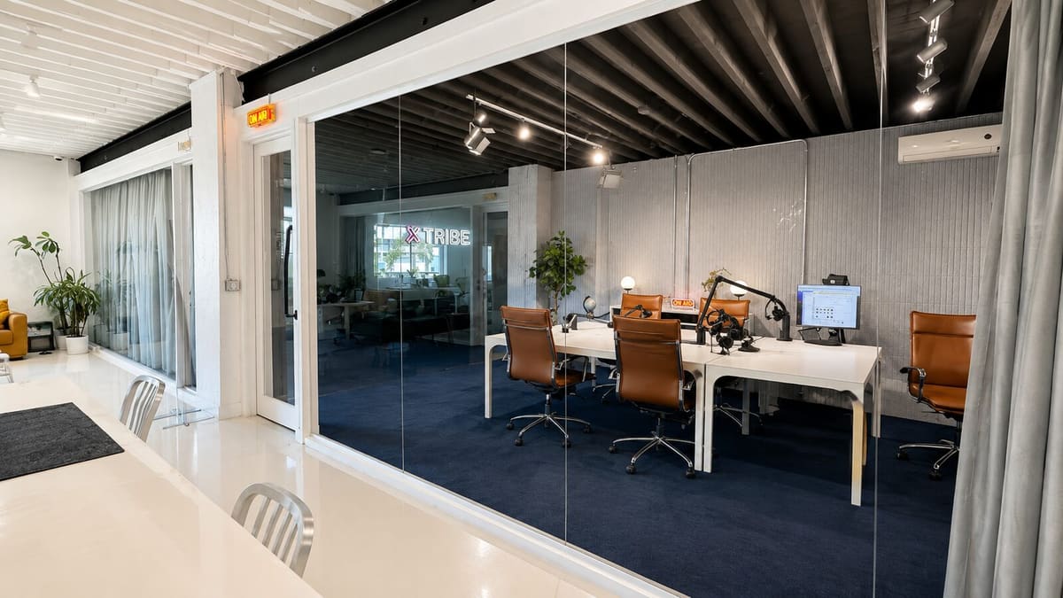 Glass-walled podcast studio with a white desk, microphones on boom arms, leather chairs and navy carpet